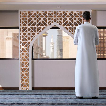 마카렘 아지아드 마카 호텔 메카 외부 사진 The photo depicts a person standing with their back facing the viewer, wearing a traditional white garment. They are in a spacious interior setting that features an ornate archway with intricate patterns, typical of Islamic architecture. Through the 
