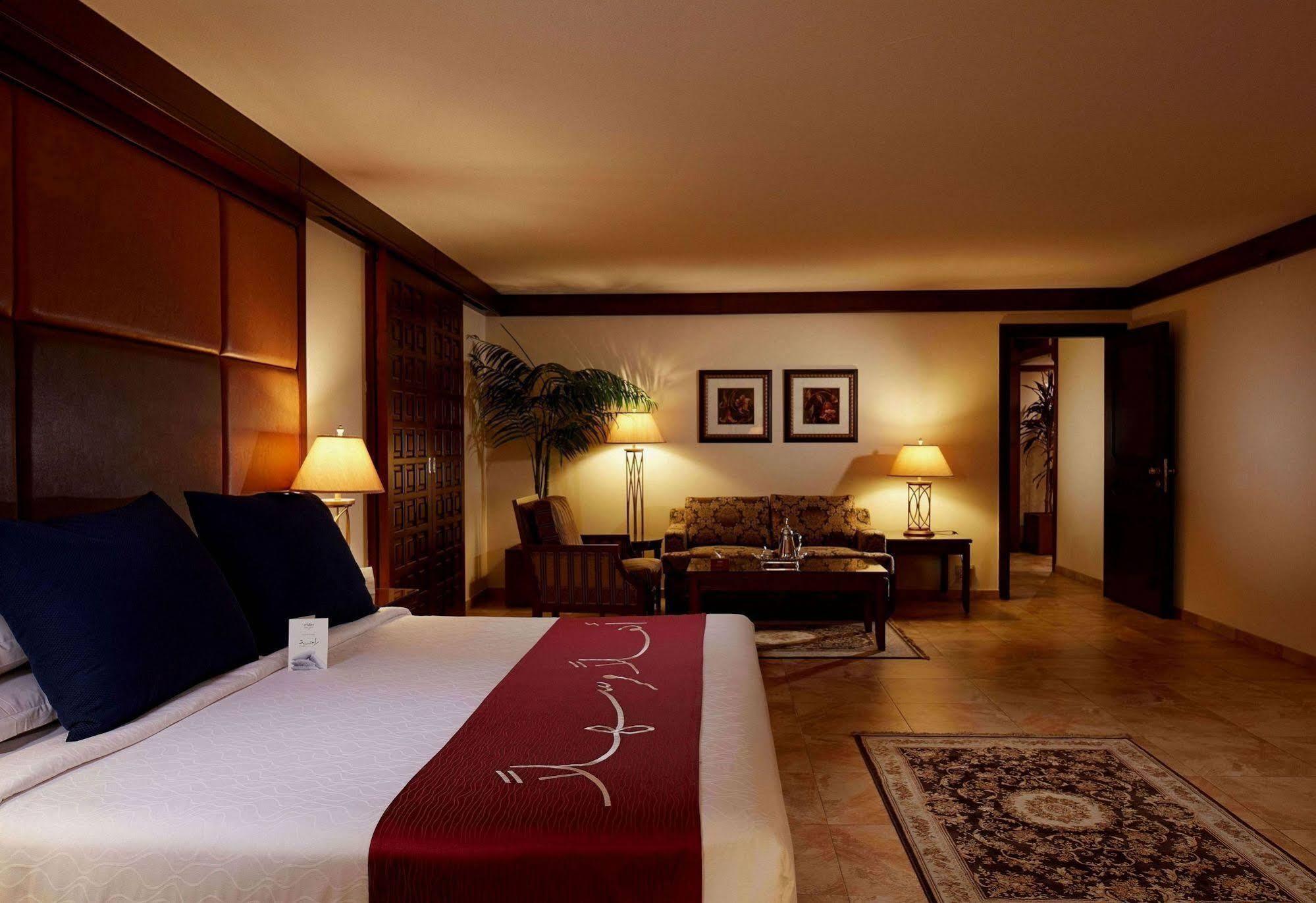 마카렘 아지아드 마카 호텔 메카 외부 사진 The photo depicts a cozy hotel room with a warm and inviting atmosphere. It features a large bed with dark blue pillows and a maroon throw blanket. On either side of the bed, there are lamps providing soft lighting. In the background, a small seating