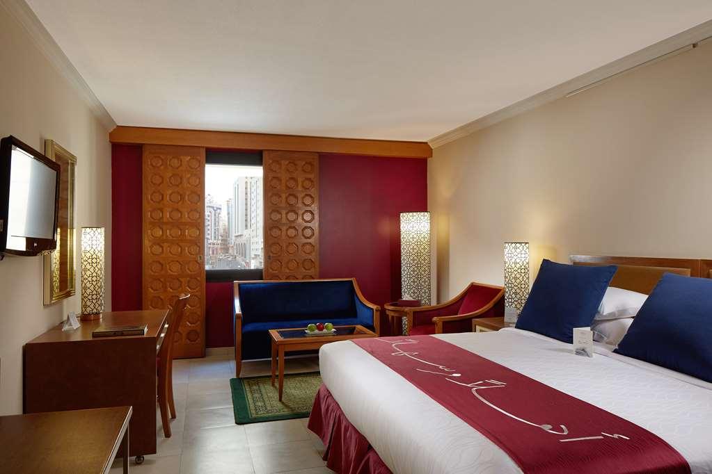 마카렘 아지아드 마카 호텔 메카 객실 사진 The photo showcases a hotel room featuring a modern design. In the center, there's a large bed with white linens and a decorative red runner. On either side of the bed, there are wooden nightstands with lamps. A blue sitting area is visible, composed