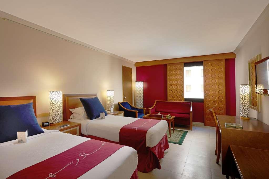 마카렘 아지아드 마카 호텔 메카 객실 사진 The photo shows a hotel room featuring two twin beds with red and white bedding. There are decorative lamps on either side of the beds. A small seating area is visible, including a blue chair and a red sofa. There is a wooden desk against one wall, a
