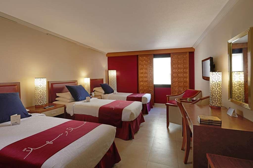 마카렘 아지아드 마카 호텔 메카 객실 사진 The photo shows a hotel room featuring three beds arranged with red and white bedding. The walls are painted in a neutral tone, and there are decorative lamps on nightstands beside each bed, providing ambient lighting. One of the walls has a window c