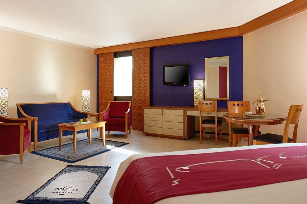 마카렘 아지아드 마카 호텔 메카 객실 사진 The image shows a hotel room with a modern, comfortable design. The room features a large bed with a red and white bedspread, a seating area that includes a blue sofa, two red chairs, and a wooden coffee table. There's a flat-screen TV mounted on the