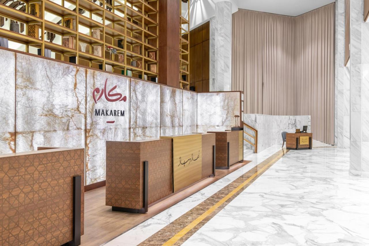 마카렘 아지아드 마카 호텔 메카 외부 사진 The photo shows a modern hotel reception area with a sleek design. The reception desk is made of a combination of wood and stone, featuring a warm color palette. The backdrop is adorned with a marble finish, giving a luxurious feel. Above the desk, t