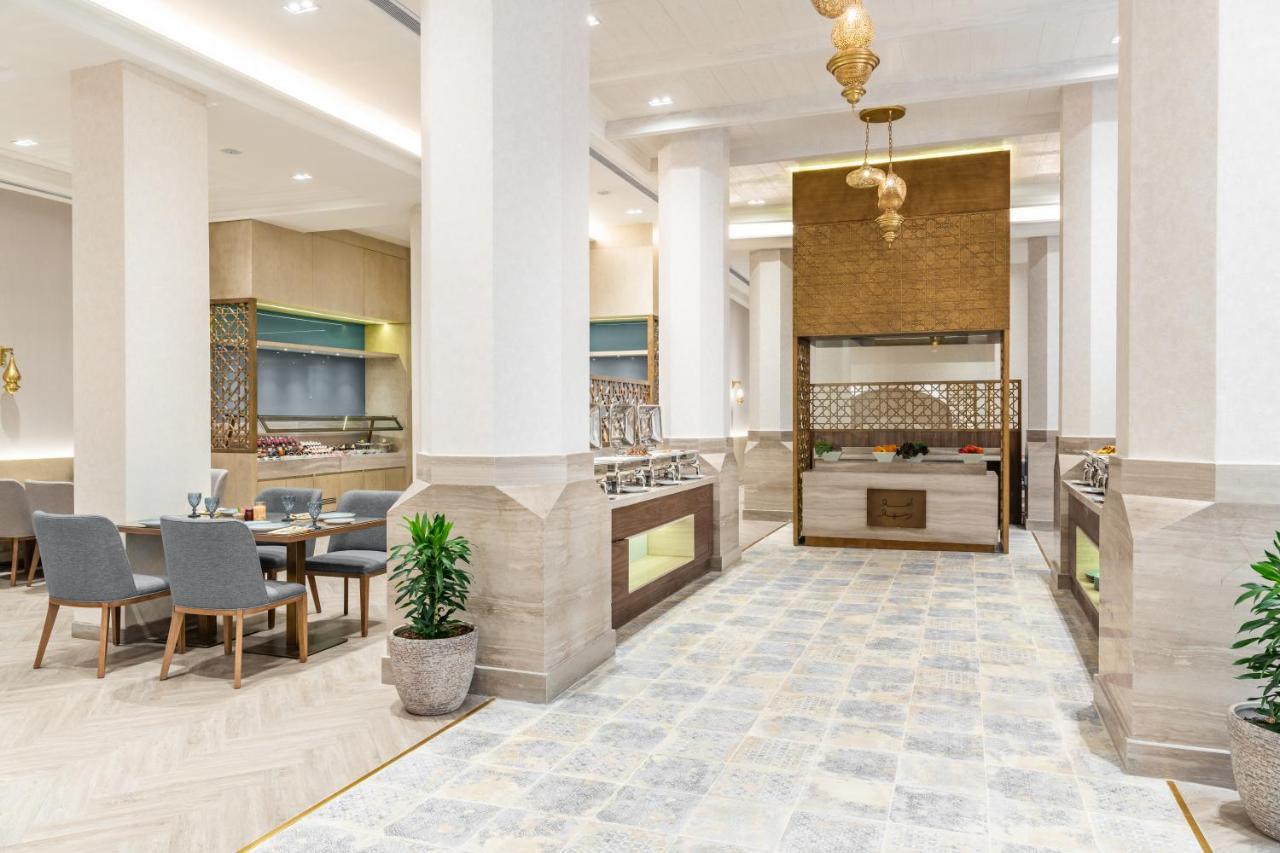 마카렘 아지아드 마카 호텔 메카 외부 사진 The photo depicts a modern, elegant dining area, likely inside a restaurant or hotel. It features a spacious layout with tall ceilings and large columns. The flooring is a light-colored tiled design. 

To one side, there are several dining tables and