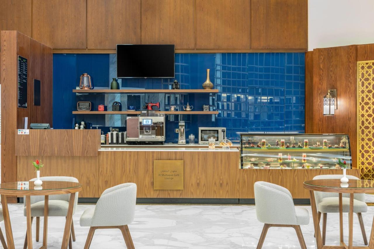 마카렘 아지아드 마카 호텔 메카 외부 사진 The photo shows the interior of a café. The design features warm wood paneling and a striking blue tiled wall. In the foreground, there are several light-colored chairs and small tables arranged neatly. Behind the counter, there's a variety of coffee