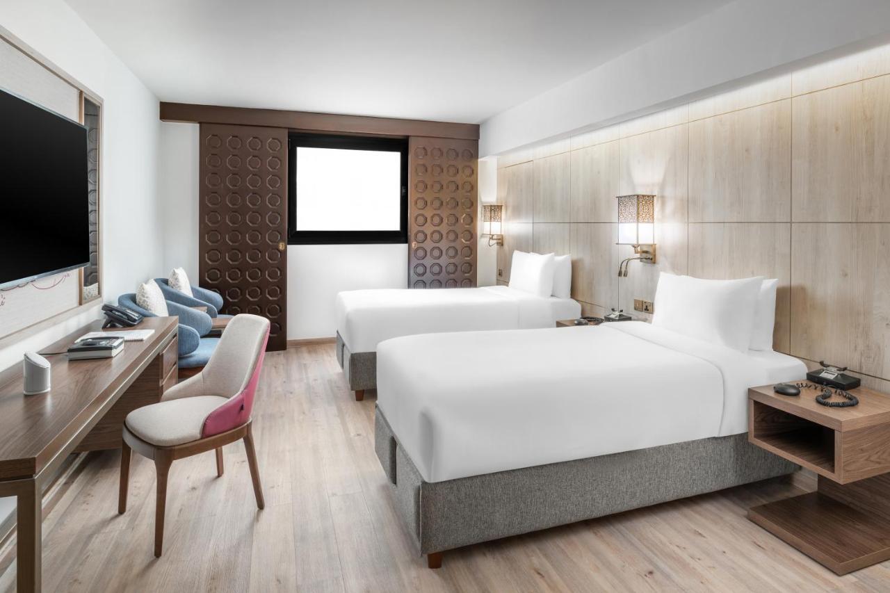마카렘 아지아드 마카 호텔 메카 외부 사진 The photo shows a modern hotel room featuring two beds with white linens. There is a wooden accent wall with a window above it. Opposite the beds, a wall-mounted television is visible. A stylish wooden desk with a chair is situated on one side of the