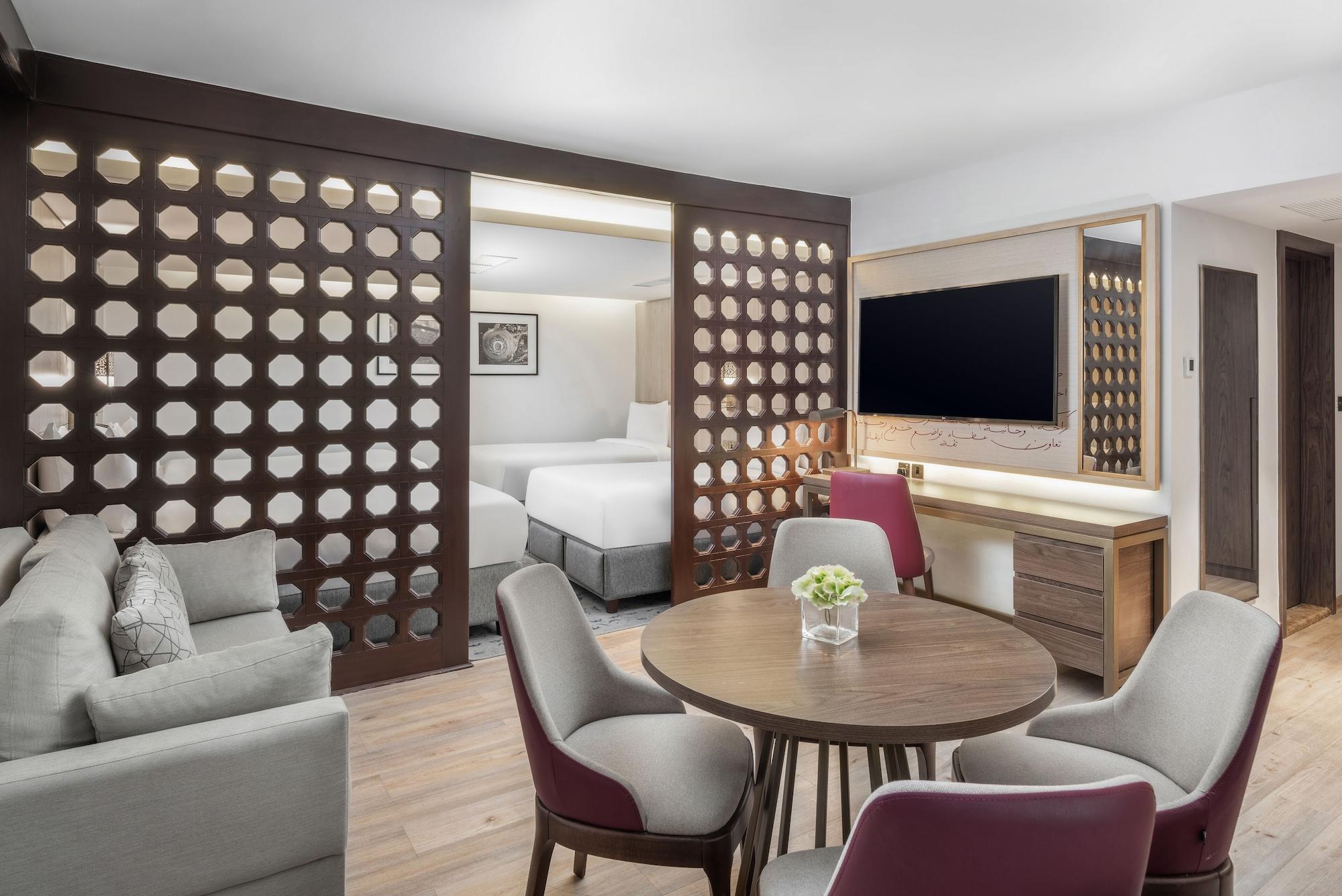 마카렘 아지아드 마카 호텔 메카 외부 사진 The photo shows a modern hotel room or suite. It features a living area with a comfortable gray sofa and a round wooden dining table surrounded by upholstered chairs. A large flat-screen TV is mounted on the wall opposite the seating area. There's a 