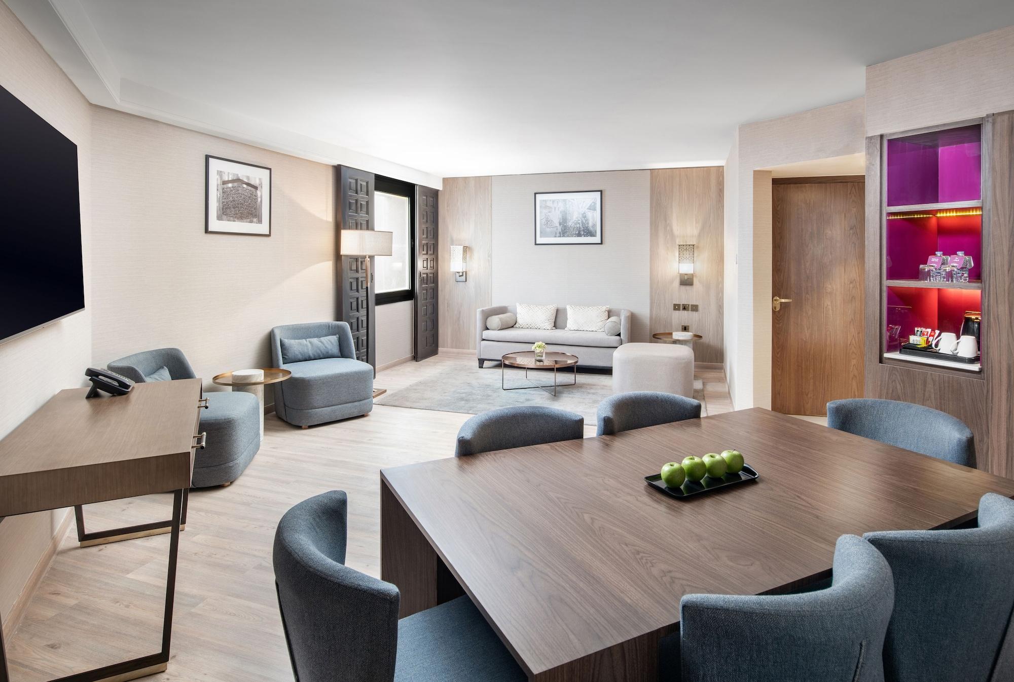 마카렘 아지아드 마카 호텔 메카 외부 사진 The photo shows a modern and elegantly furnished living area. It features a spacious layout with a comfortable seating arrangement that includes a sofa and armchairs. There is a large wooden dining table with several chairs around it. The walls are a