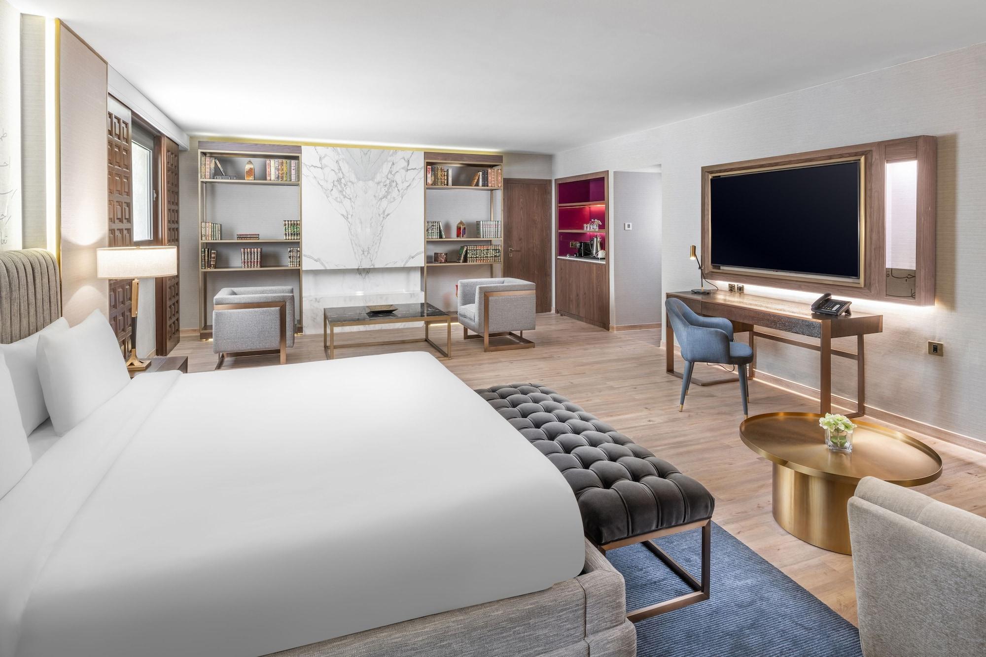 마카렘 아지아드 마카 호텔 메카 외부 사진 The photo shows a modern hotel room designed with a contemporary aesthetic. The room features a large bed with white bedding, complemented by a stylish tufted bench at the foot of the bed. To the side, there are two comfortable armchairs positioned a