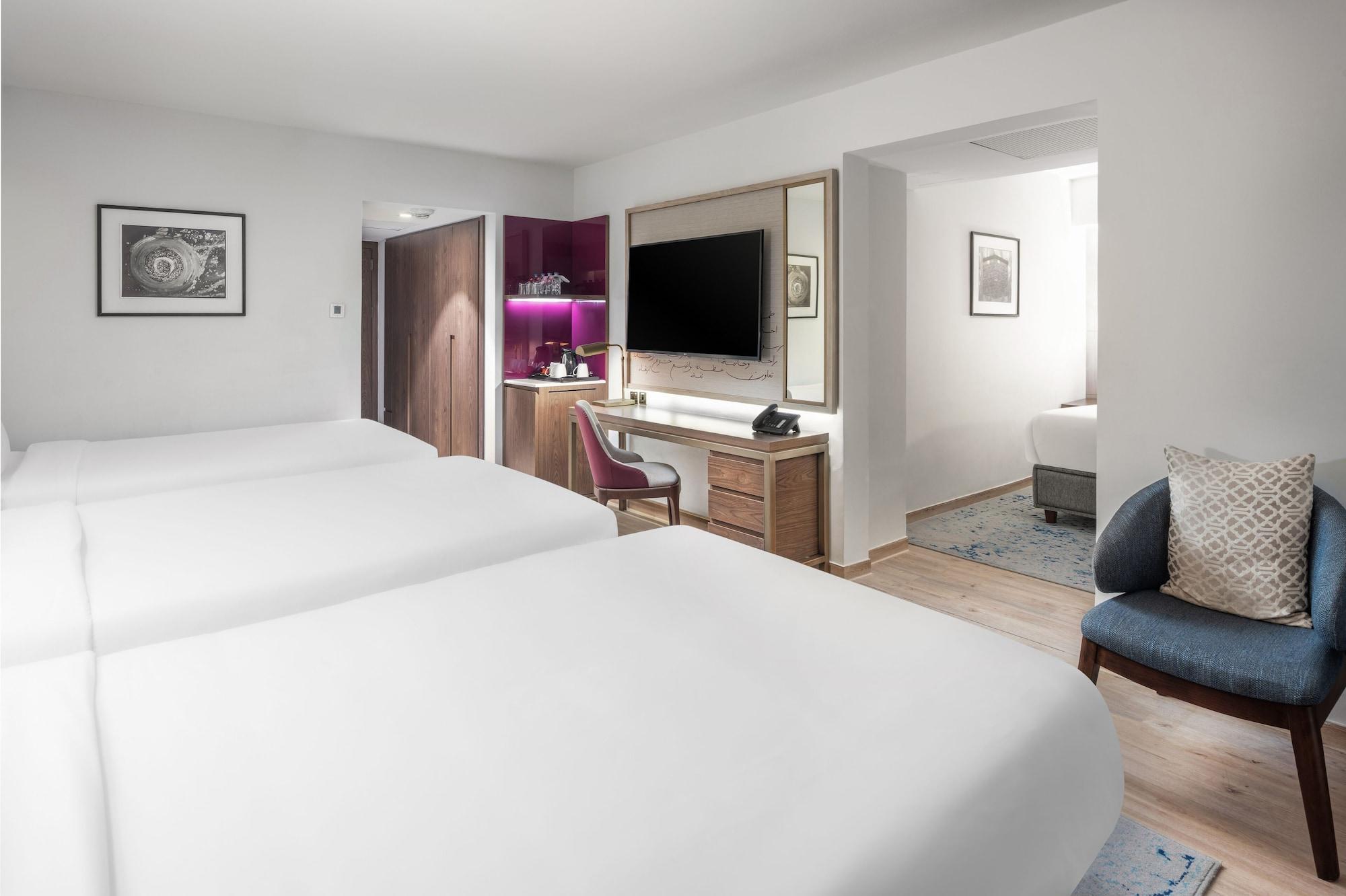 마카렘 아지아드 마카 호텔 메카 외부 사진 The photo shows a modern hotel room featuring two white beds with smooth linens. The room has a minimalistic design with light-colored walls and wooden flooring. On one side, there is a desk and chair next to a television mounted on the wall, along w