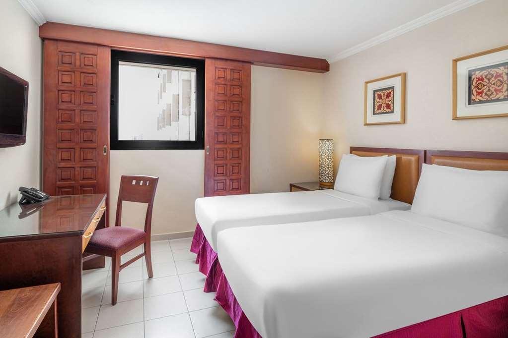 마카렘 아지아드 마카 호텔 메카 객실 사진 The photo shows a hotel room with two beds, each covered with white bedding. There is a wooden desk and a chair near the beds. The room features a window with a wooden frame and a decorative lamp beside the beds. On the wall, there are two framed art