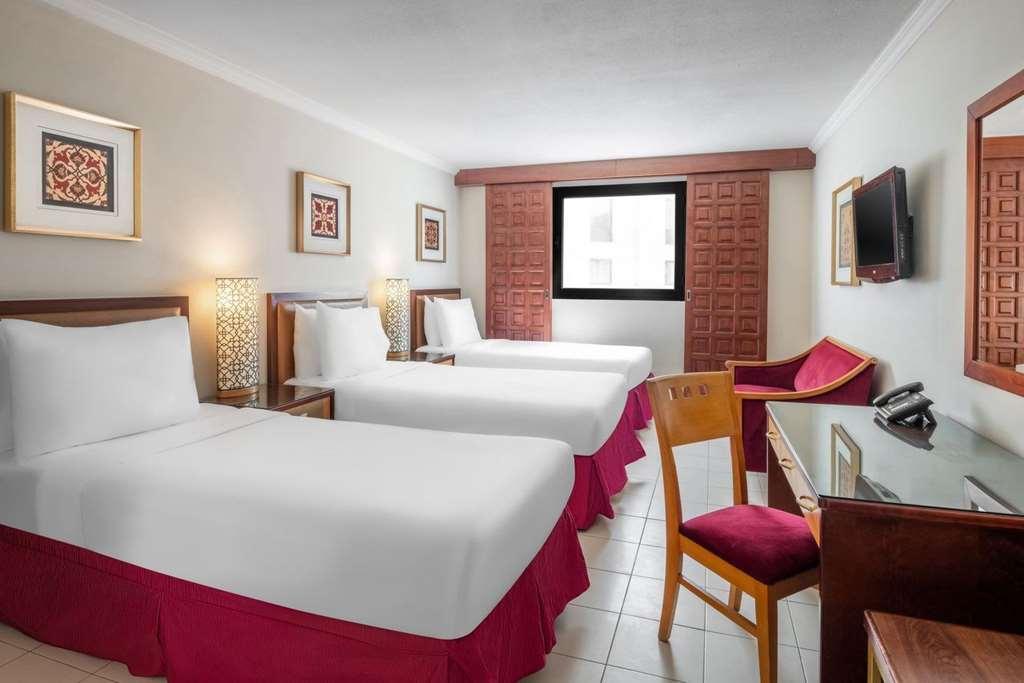 마카렘 아지아드 마카 호텔 메카 객실 사진 The photo shows a hotel room featuring two twin beds with white linens and red bed skirts. There are decorative elements on the walls, including framed artwork. A large window provides natural light, while a desk and a chair are positioned next to it