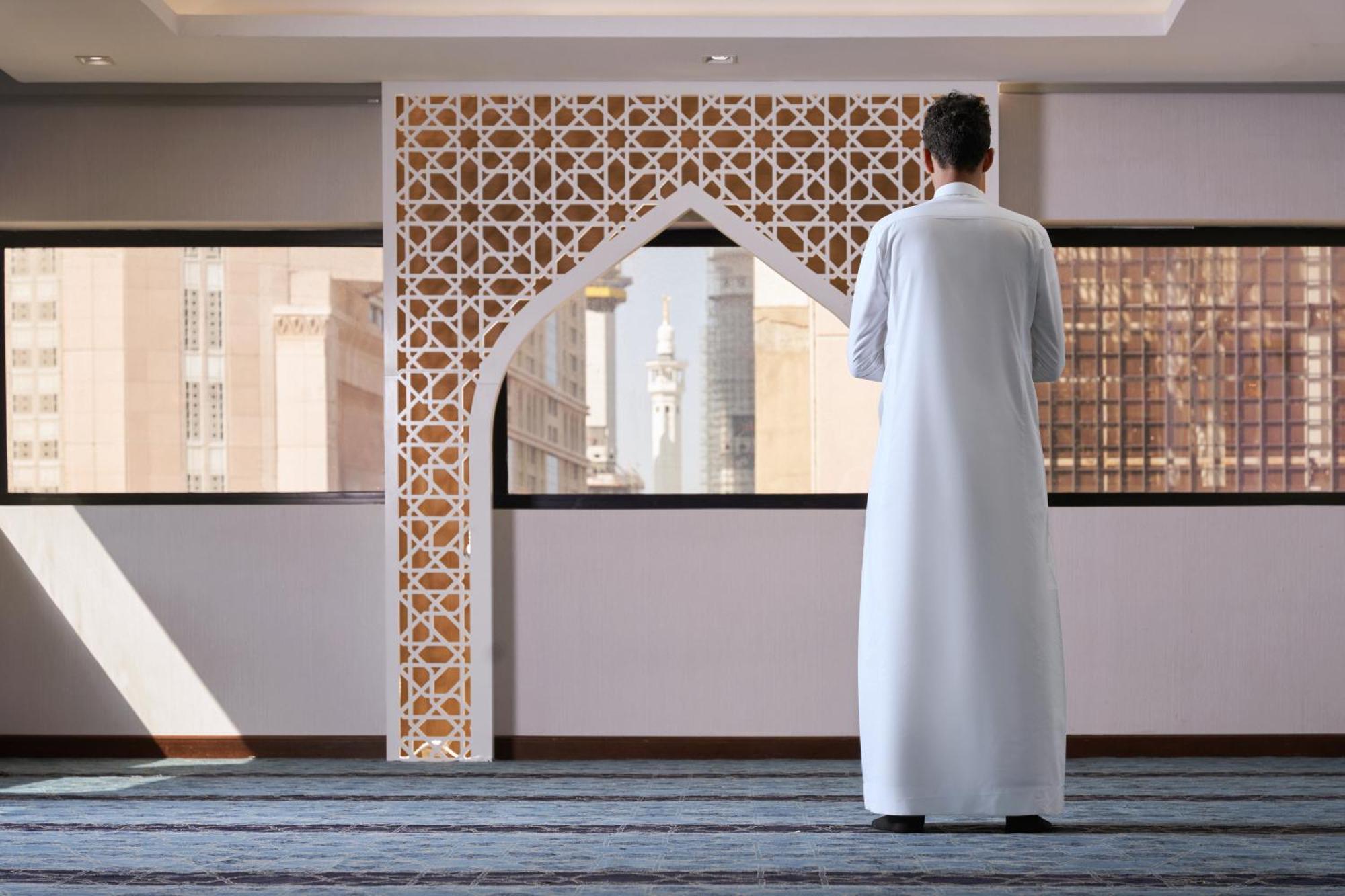 마카렘 아지아드 마카 호텔 메카 외부 사진 The photo depicts a person standing with their back facing the viewer, wearing a traditional white garment. They are in a spacious interior setting that features an ornate archway with intricate patterns, typical of Islamic architecture. Through the 