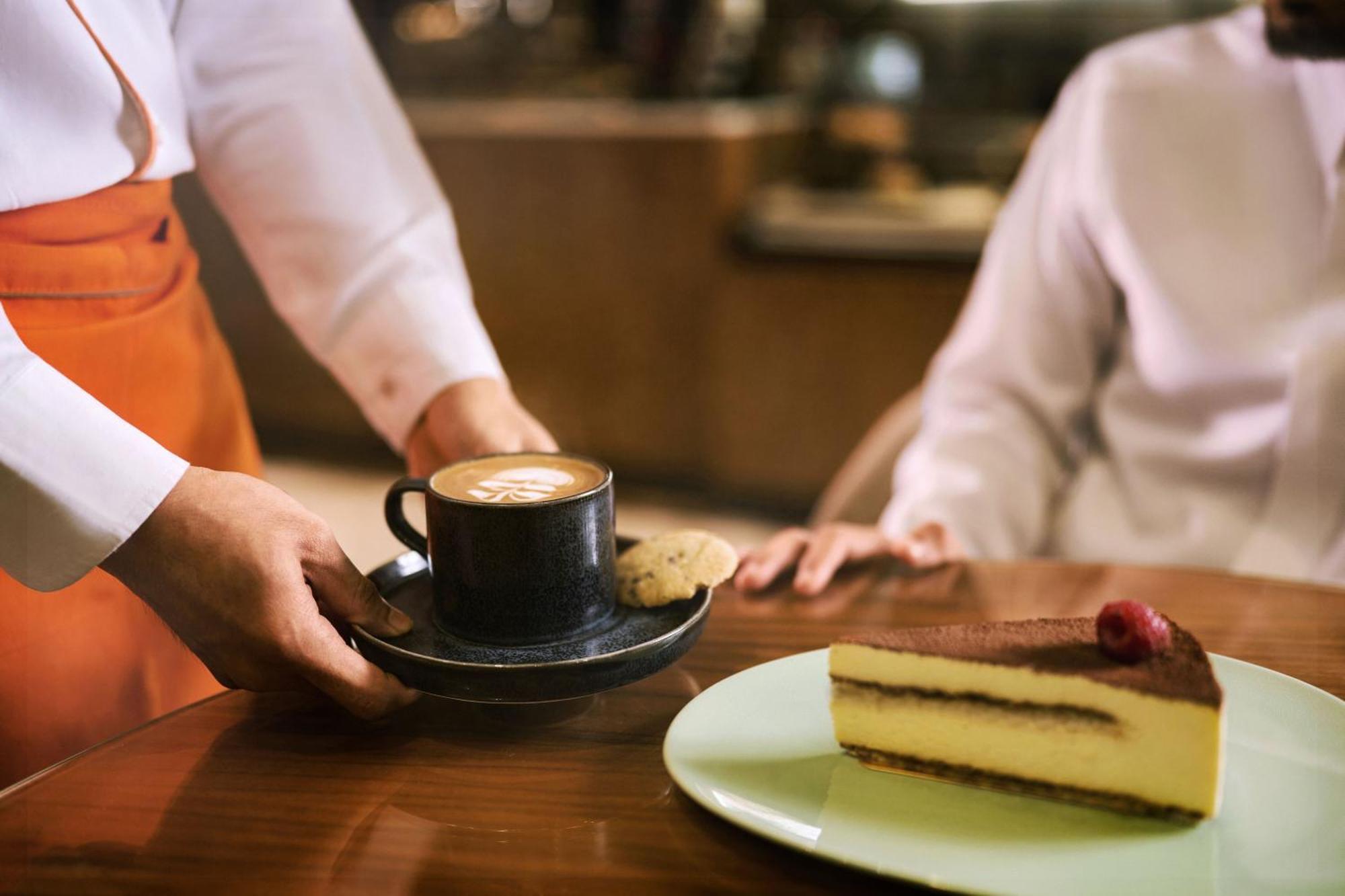 마카렘 아지아드 마카 호텔 메카 외부 사진 The photo shows a hand presenting a cup of coffee on a saucer, accompanied by a cookie. In the foreground, there is a slice of cake on a light green plate, garnished with a raspberry. The setting appears to be a café or restaurant, with a blurred bac