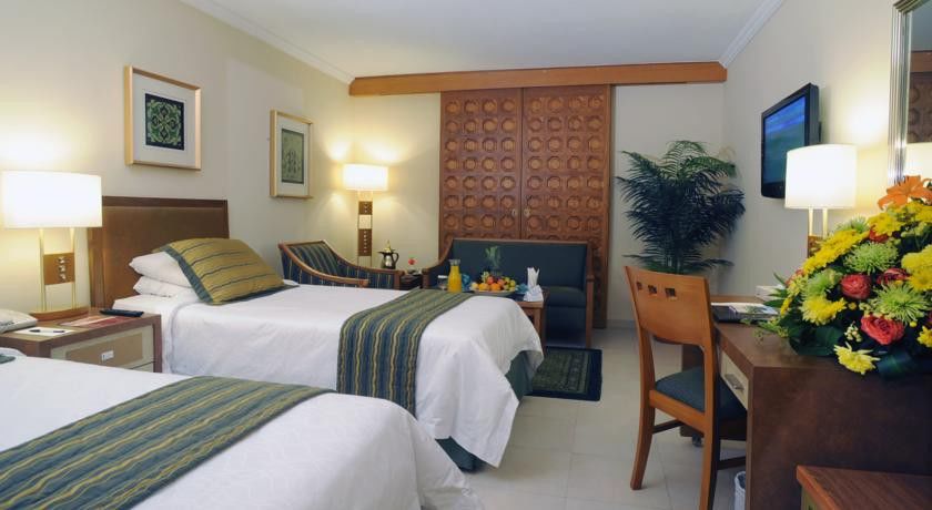 마카렘 아지아드 마카 호텔 메카 외부 사진 The photo shows a hotel room featuring two single beds with white linens and green and blue striped throw blankets. There is a wooden headboard behind the beds and a decorative wooden partition or wall in the background. A cozy seating area is visibl