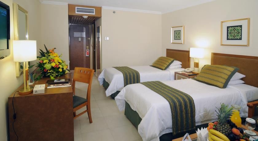 마카렘 아지아드 마카 호텔 메카 외부 사진 The photo shows an interior of a hotel room featuring two twin beds with white linens and green decorative accents. The walls are painted in a light beige color, and there is a small table next to one of the beds. A desk with a chair is positioned ne
