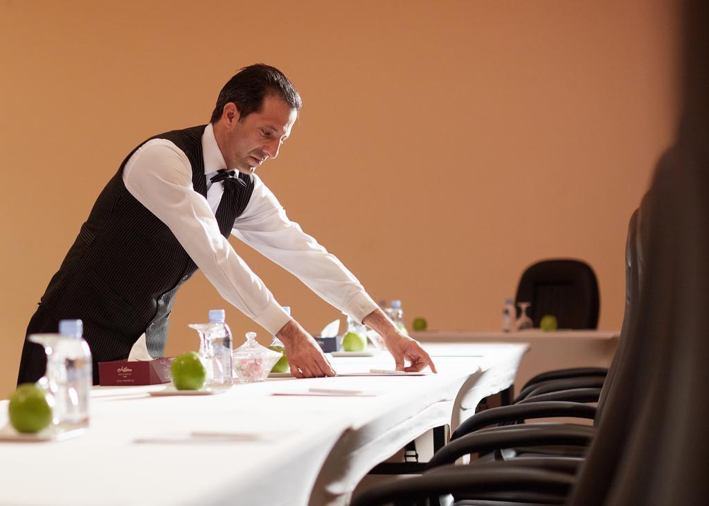마카렘 아지아드 마카 호텔 메카 외부 사진 The image shows a waiter or service staff member in a formal attire, which includes a vest and bow tie. He is preparing a long table, likely for an event or meeting. The table is set with items such as water bottles and green apples, and there are se